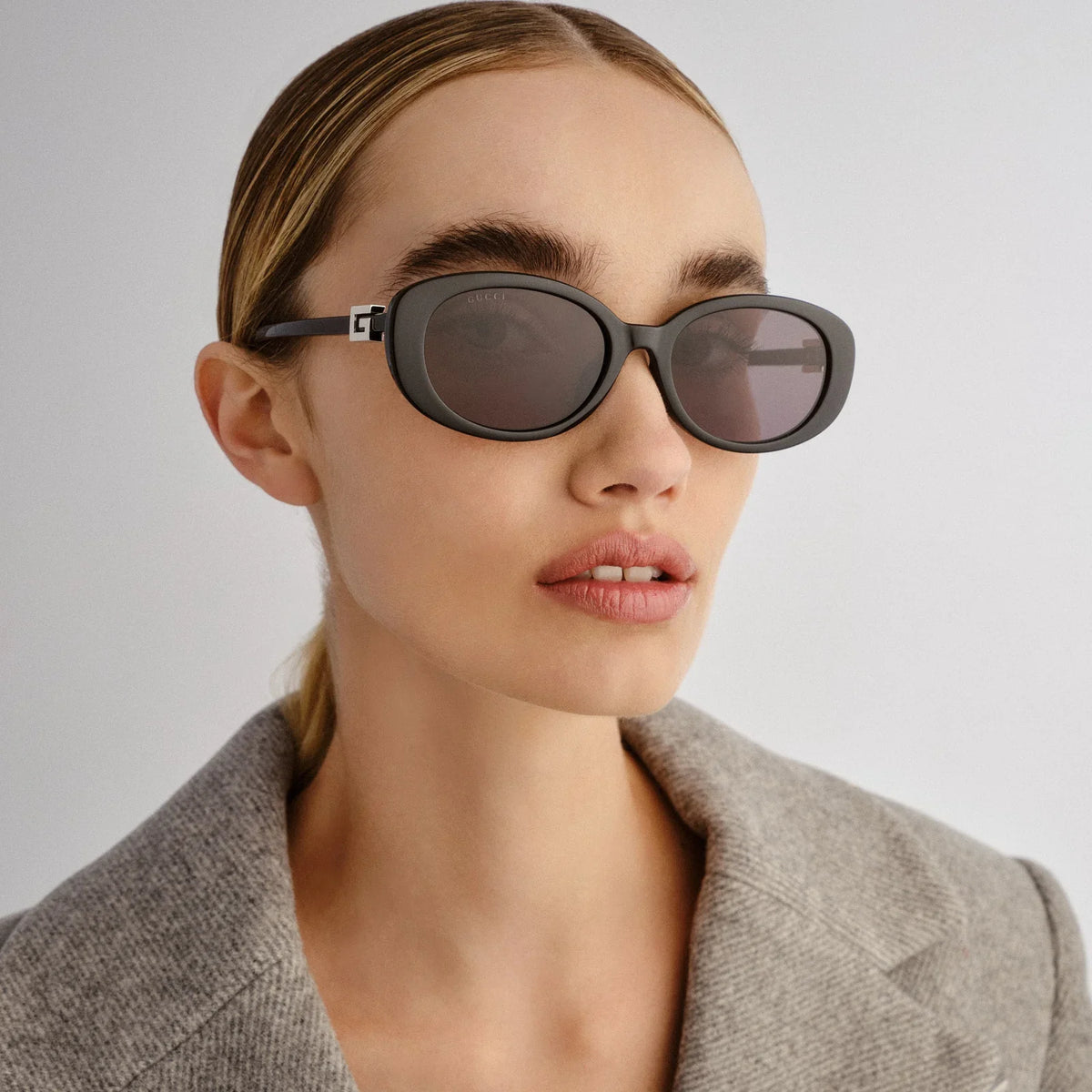 Woman wearing oversized dark grey oval sunglasses and a grey blazer.